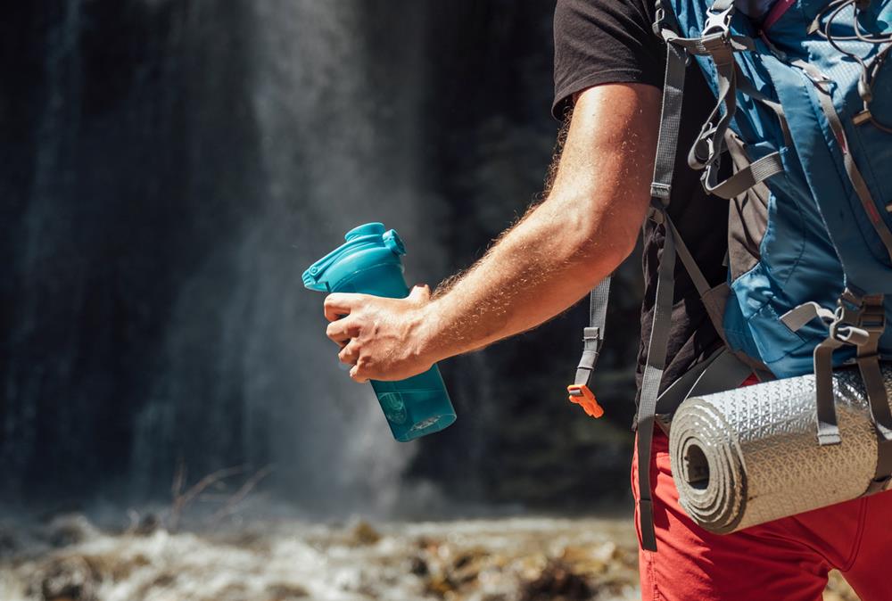 Man dressed in hiking gear standing near waterfall with hand holding water bottle