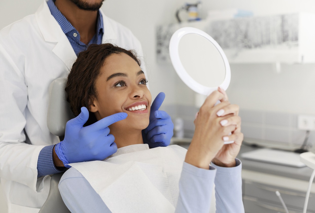 Attractive black lady checking her beautiful smile in mirror after stomatological treatment